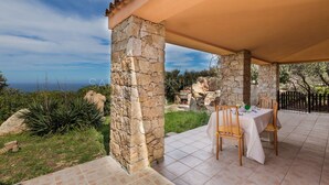 Un espace extérieur abrité, idéal pour prendre ses repas, avec des piliers en pierre, une table dressée pour deux personnes et une vue sur la mer.