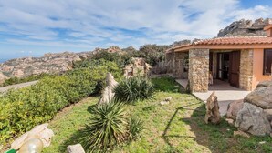 Une maison en pierre avec un toit de tuiles, entourée d’un jardin agrémenté de rochers et de plantes, et offrant une vue sur les collines en arrière-plan.