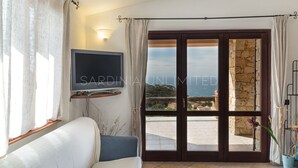 Une pièce comprenant un canapé, un téléviseur à écran plat fixé au mur, une grande porte-fenêtre coulissante donnant sur un espace extérieur et une plante en pot.