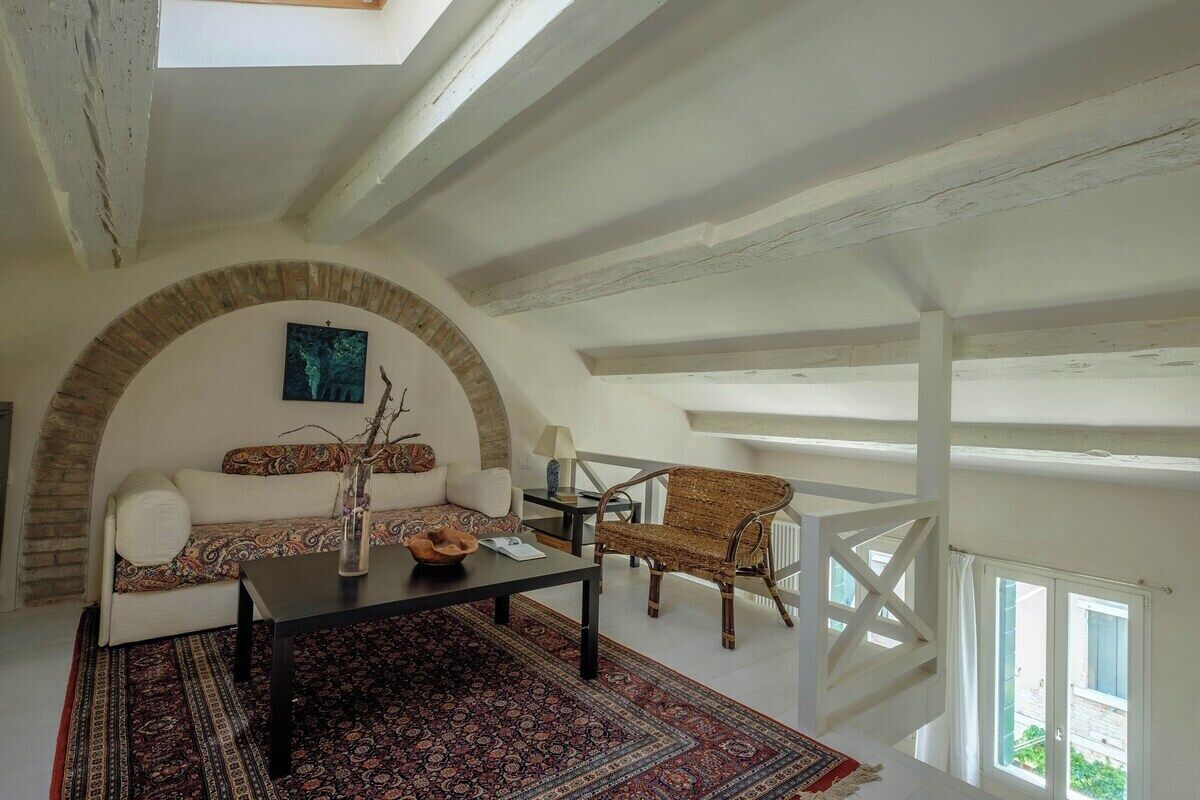 Una stanza in stile loft con soffitto alto, travi a vista, un arco in mattoni, un divano, una sedia, un tavolo e vista da una finestra.