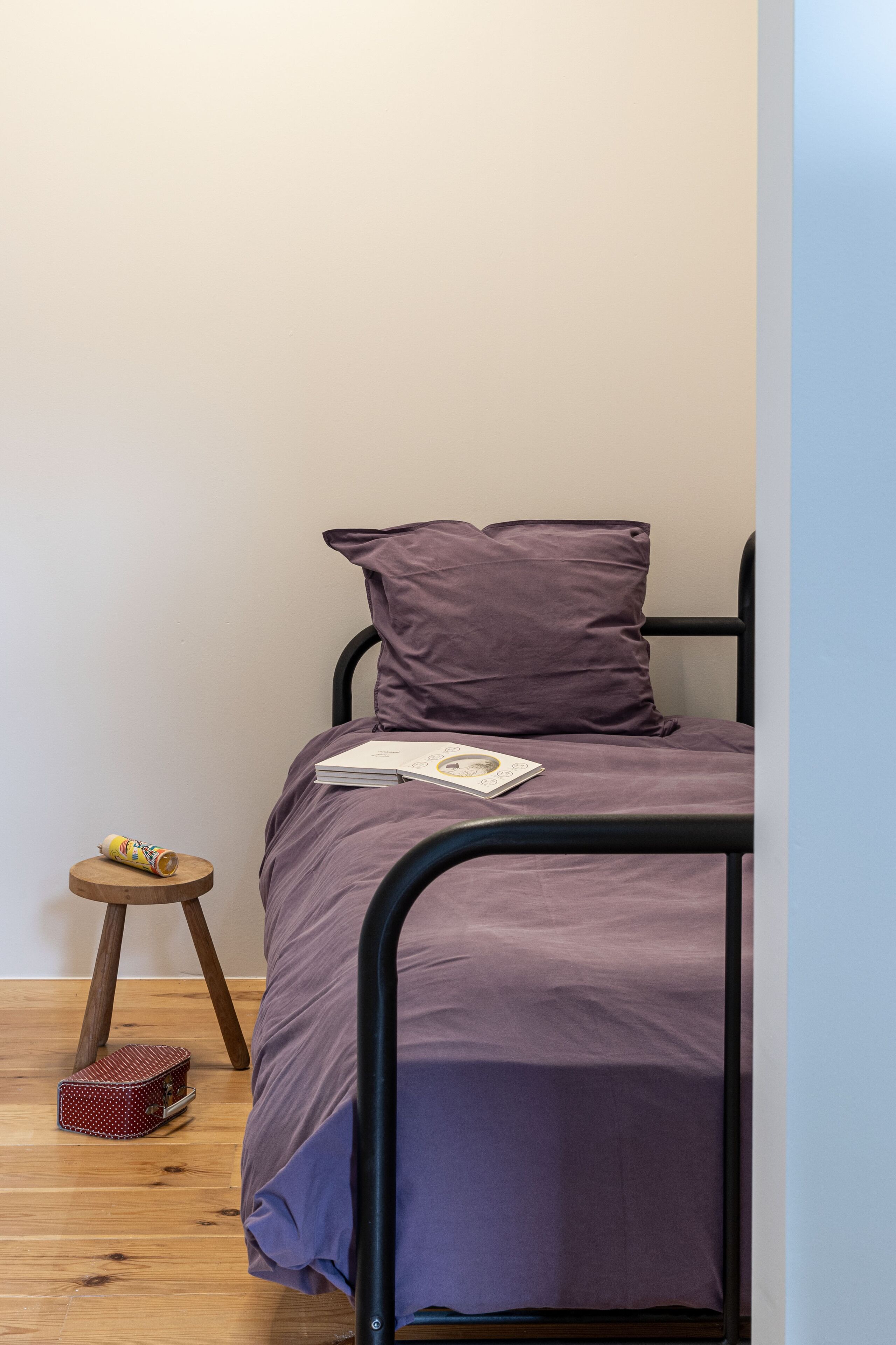 Un lit simple avec une couvre-lit et des oreillers violets, un petit tabouret en bois avec une boîte à motifs à pois et une boîte de conserve à côté, et une fenêtre donnant sur le ciel.