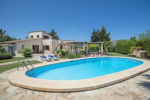 Une piscine avec une terrasse en pierre, un patio couvert doté de chaises longues, et une maison avec un toit en tuiles en arrière-plan.