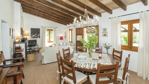Un coin repas avec une table en bois dressée pour quatre personnes, entourée de chaises en bois avec des coussins blancs. Un lustre est suspendu au plafond, et on aperçoit un téléviseur à l’arrière-plan.