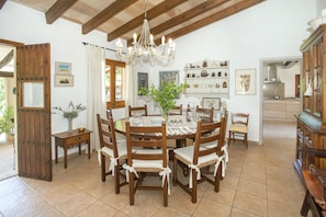 Une salle à manger avec une table et des chaises en bois, un lustre et une cuisine visible à travers une porte ouverte.