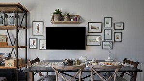 Un coin repas avec une table dressée pour deux personnes, entouré de chaises en bois dotées d’assises rembourrées. Un téléviseur est fixé au mur au-dessus de la table, et des tableaux encadrés ornent le mur.