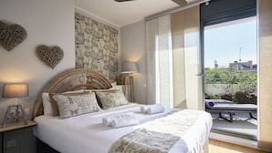 Une chambre avec un grand lit, un ventilateur de plafond, un mur décoré d’un collage de papier, une table de chevet avec une lampe, et une vue sur l’extérieur.