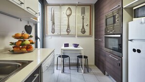 Une petite cuisine équipée d’un évier, d’une cuisinière et d’un four. On y trouve deux tabourets et une petite table avec un bol et un livre.