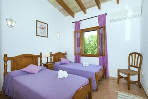 A bedroom with two wooden beds, purple bedding, a wooden chair, a nightstand with a lamp, a framed picture on the wall, and a window with purple curtains. - VILLA  BOBIS - RUSTIC STAY (Pollensa)