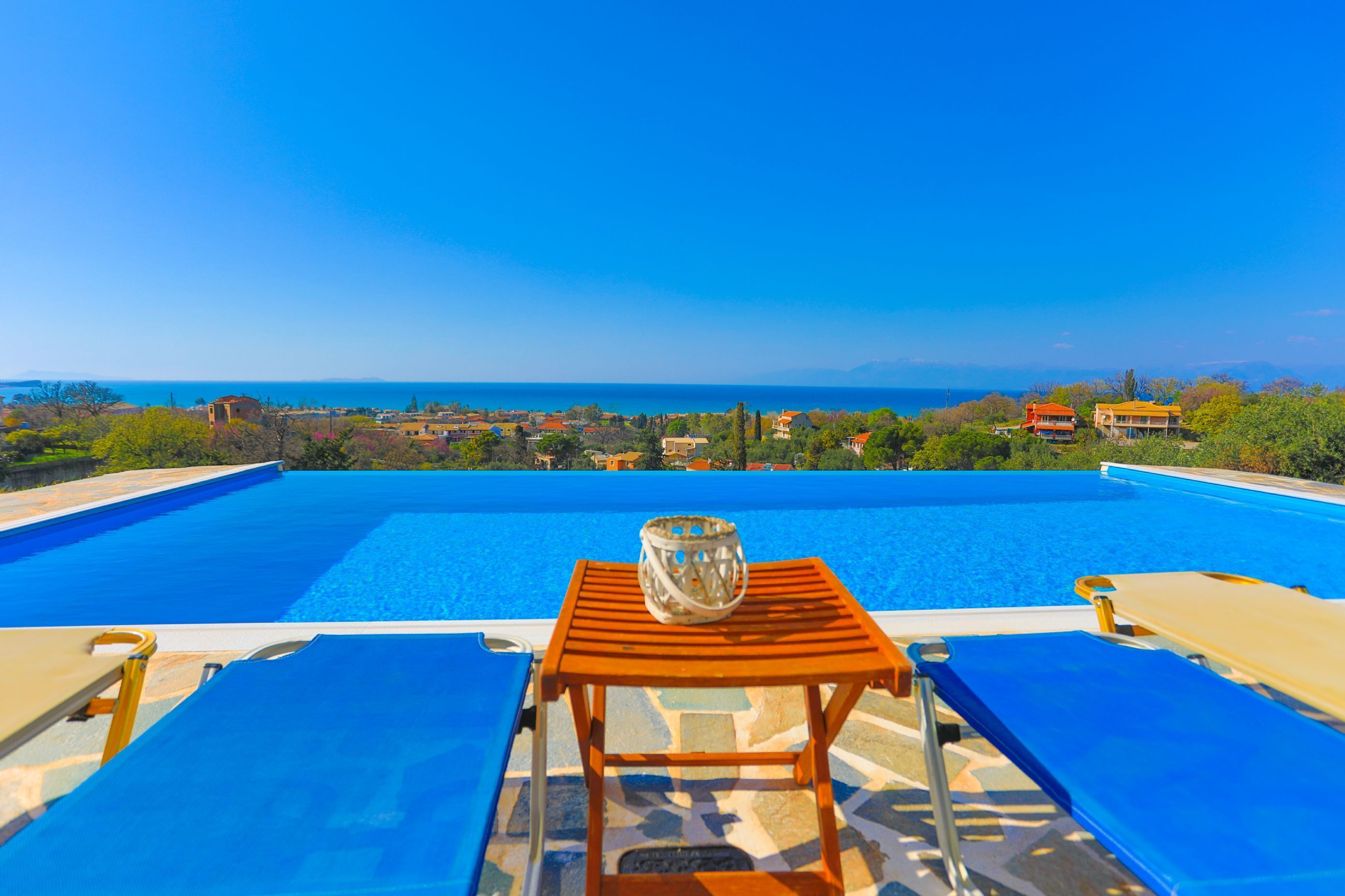 A poolside view with a wooden table and lounge chairs, overlooking a scenic landscape and the sea.