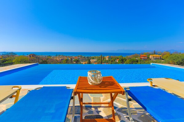 A poolside view with a wooden table and lounge chairs, overlooking a scenic landscape and the sea.