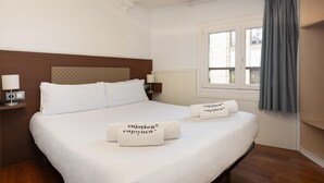 A hotel room with a large bed, two bedside tables with lamps, a window with curtains, and a view of buildings outside.