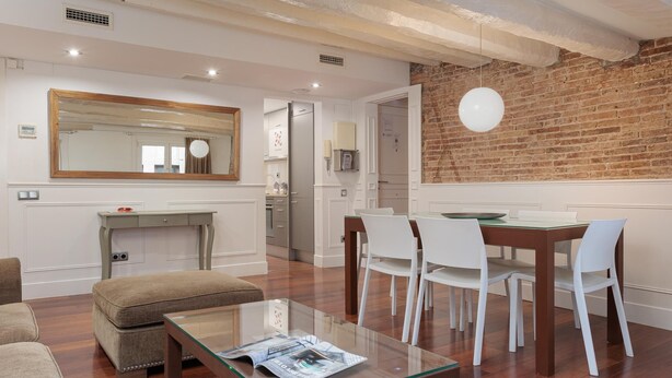 A modern living room with a glass coffee table, a dining area with white chairs, a large mirror, and a brick wall.