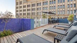 A rooftop terrace with lounge chairs, a table, and a view of a large building with a classical architectural style.