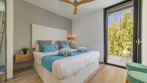 Een moderne slaapkamer met een groot bed, een plafondventilator, een nachtkastje met lamp en uitzicht op de omgeving via een glazen schuifdeur.