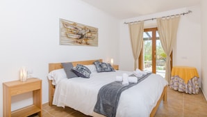 Un dormitorio ordenado con cama, mesitas de noche, un cuadro en la pared y una ventana con cortinas.