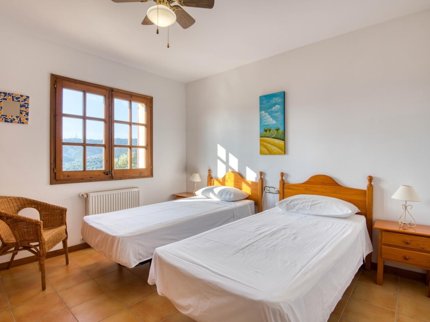A room with two single beds, a wooden headboard, a window with a view, a radiator, a wicker chair, and a painting on the wall.