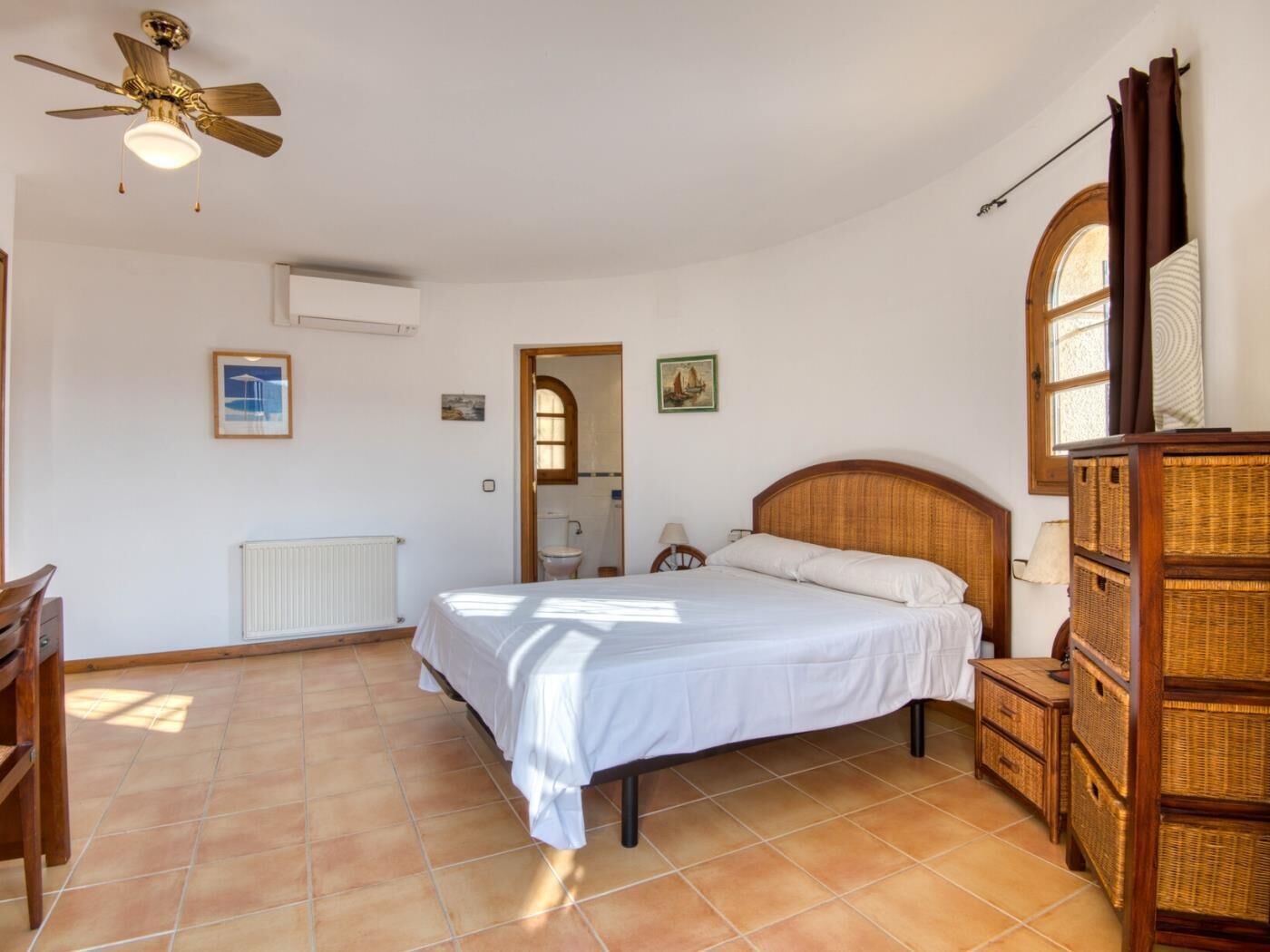 Un dormitorio con cama, mesita de noche de madera, cómoda de mimbre, ventilador de techo y ventana con cortinas.