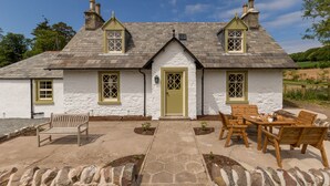 A white cottage with a green door, two chimneys, and a stone pathway leading to a wooden table and chairs.