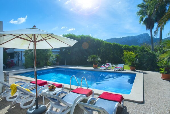 Una piscina con sillones de descanso y una mesa, rodeada de vegetación y una montaña al fondo.