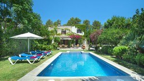 Una piscina rectangular con sillas de descanso y una sombrilla, rodeada de exuberante vegetación y una casa al fondo.