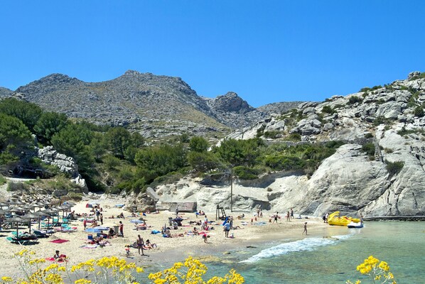A beach with clear waters, rocky cliffs, and a variety of beach umbrellas and sun loungers. - VILLA EL CAMPET - RUSTIC STAY (Pollensa)