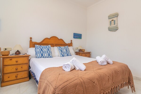 Una cama bien tendida con un edredón marrón, dos almohadas y dos toallas enrolladas sobre una mesita de noche de madera.