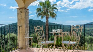 A balcony with a table and chairs, a palm tree, and a view of green hills. - Villa Bunganvilla, Calonge - Costa Brava (Calonge)