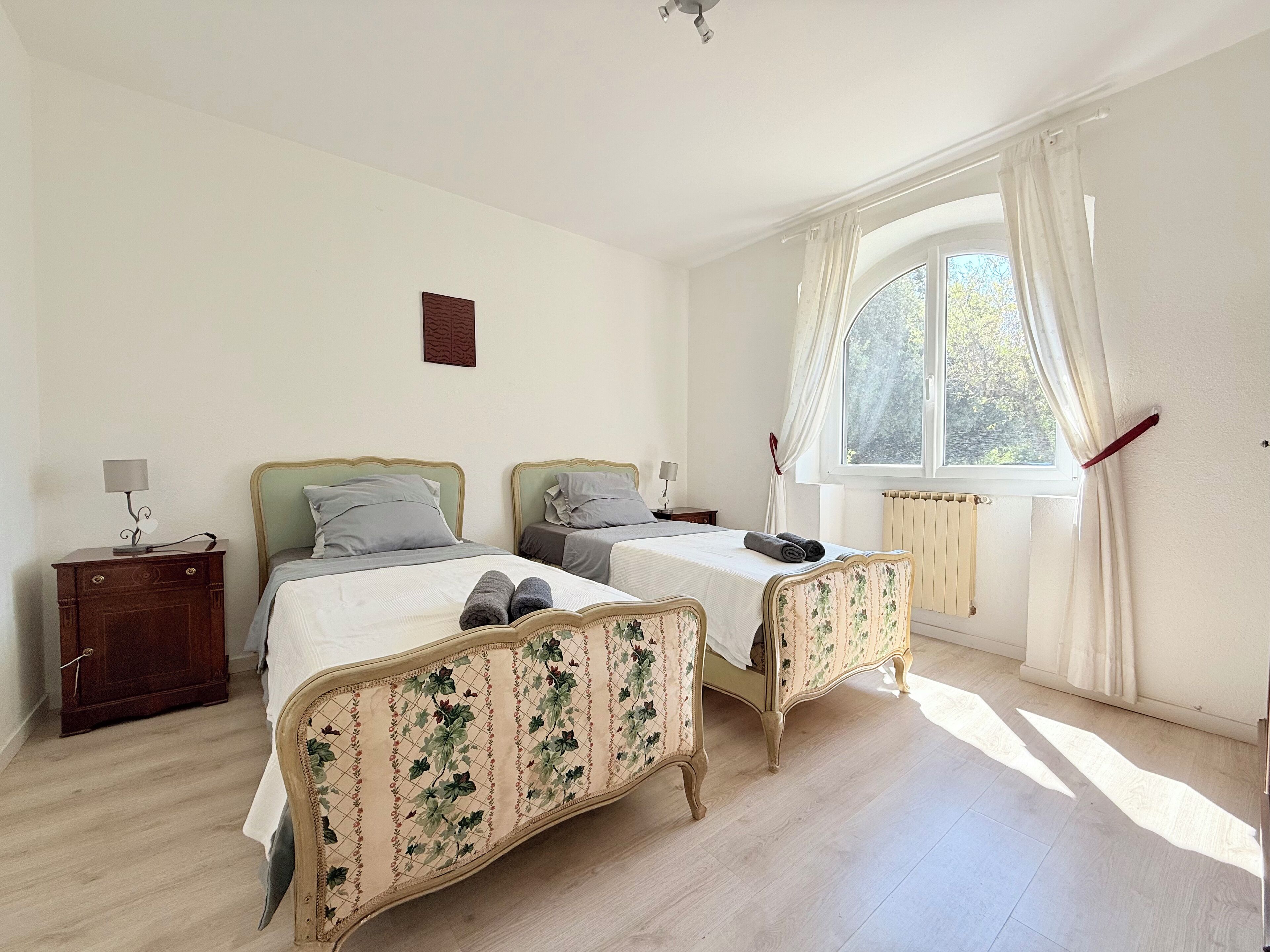 A bedroom with two beds, a nightstand, a window with curtains, and a radiator.