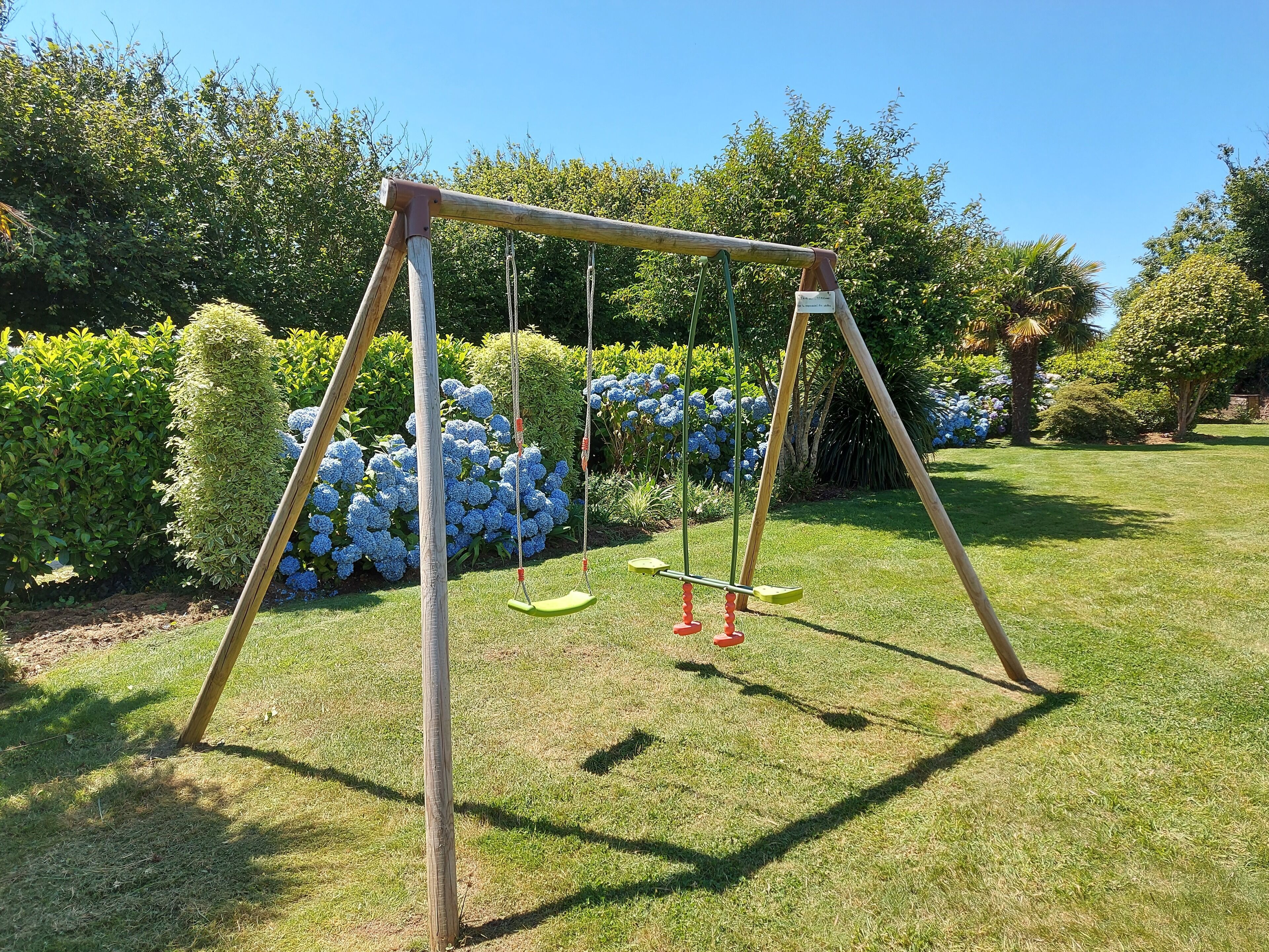 Un conjunto de columpios de madera en un jardín bien cuidado, con abundante vegetación y arbustos florecientes.
