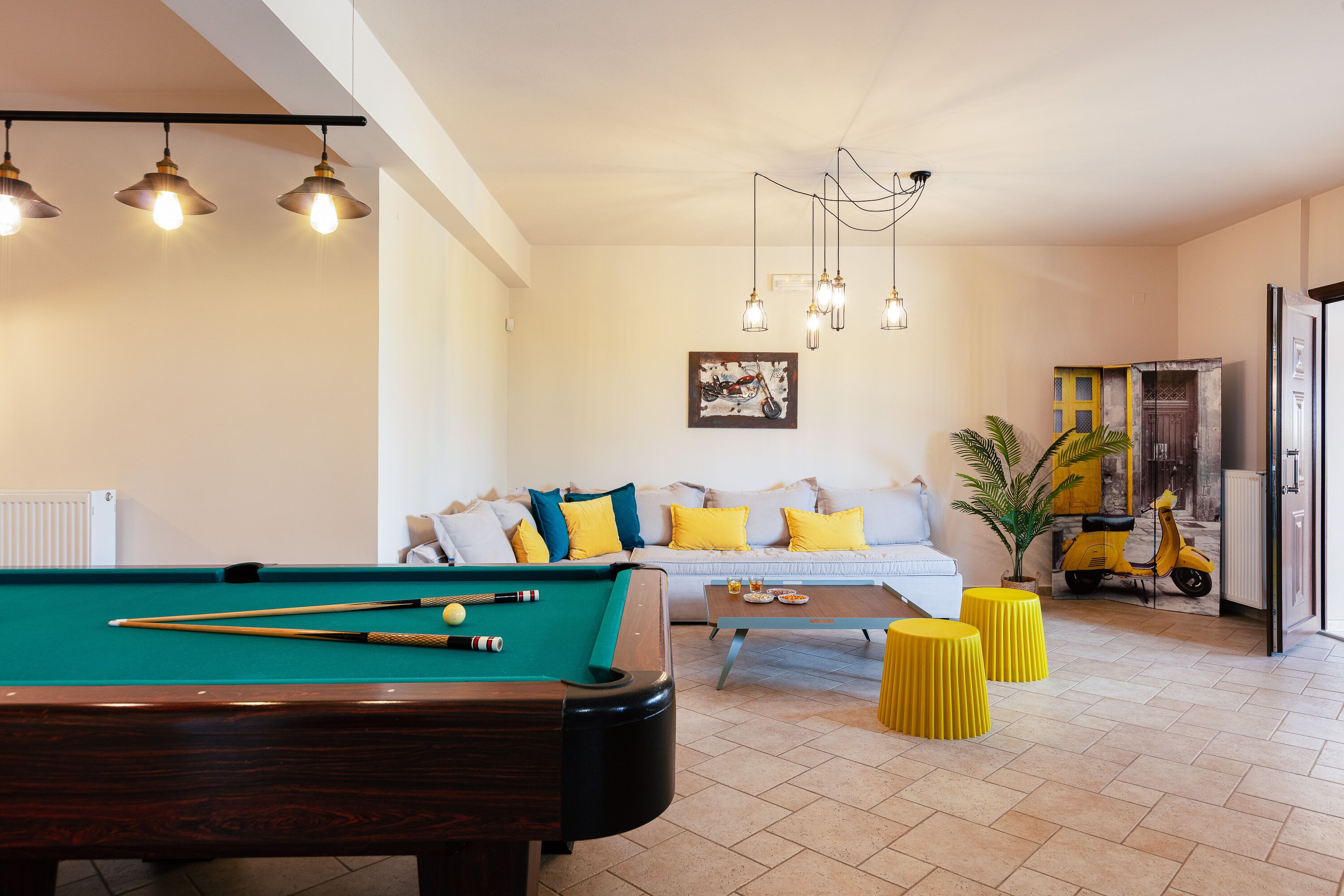 A pool table with cues, a sofa with yellow cushions, and a scooter in a living area with tiled flooring.