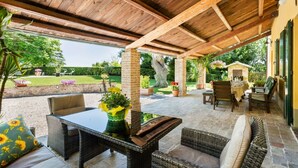 An outdoor patio area with wicker furniture, a table with a vase of flowers, and a view of a garden and a building in the background. - Villa delle Vigne, villa with large garden and pool, close to the Adriatic coast (Pesaro)