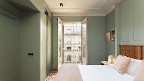 A bedroom with a bed, a nightstand, a lamp, and a view of a building through an open door. - Luxurious Oasis Paseo de Gracia (Barcelona)