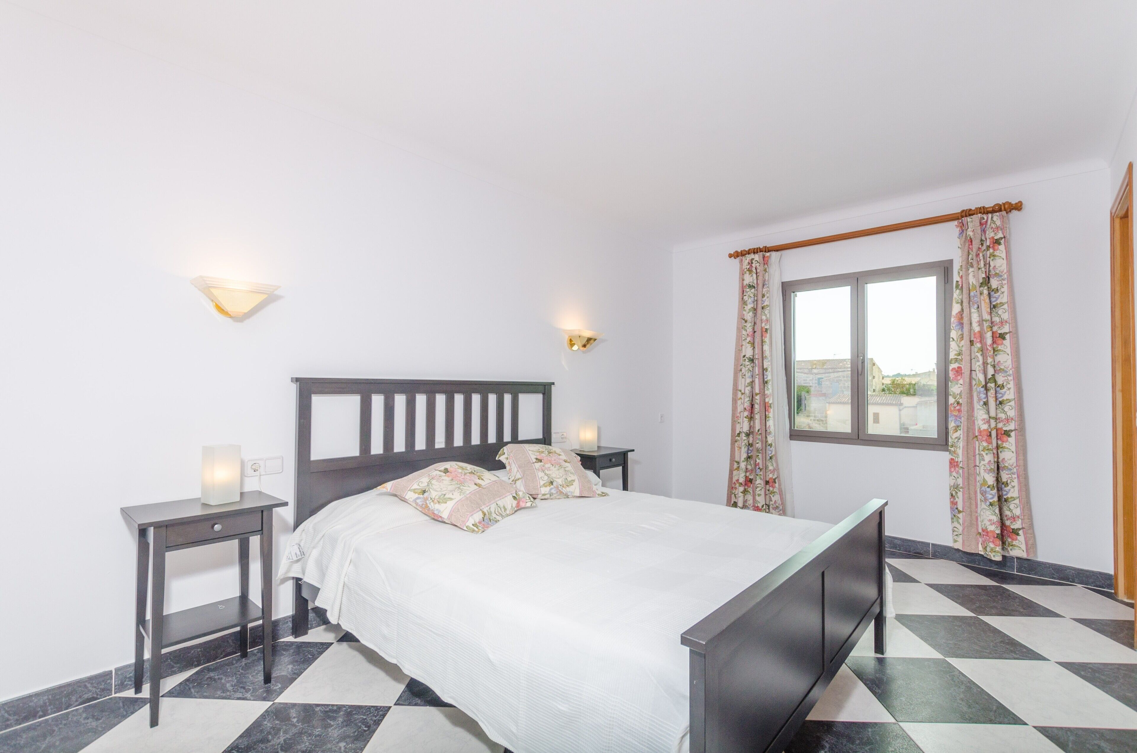 A bedroom with a bed, two bedside tables, wall-mounted lights, a window with curtains, and a checkered floor.