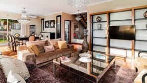 A spacious living room with a large sofa, a coffee table, and a TV mounted on a wall. There is a staircase leading to an upper level, and a dining area with chairs and a table.