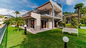 A modern two-story house with a balcony, surrounded by well-maintained grass and palm trees.