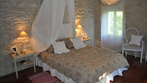 A bedroom with a stone wall, a canopy bed, bedside lamps, a chair, and a small table with a lamp.