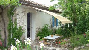 A rustic stone building with a white exterior and blue shutters, a covered patio with a table and chairs, and a garden with flowering plants.