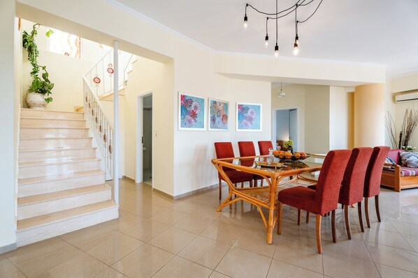 A spacious dining area with a round table and red chairs, a staircase with a potted plant, and three framed artworks on the wall.