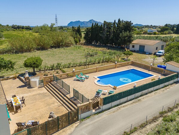 Ein rechteckiger Swimmingpool mit gefliestem Sonnendeck, umgeben von einem niedrigen Zaun. Liegestühle und ein Tisch mit Stühlen befinden sich rund um den Pool.