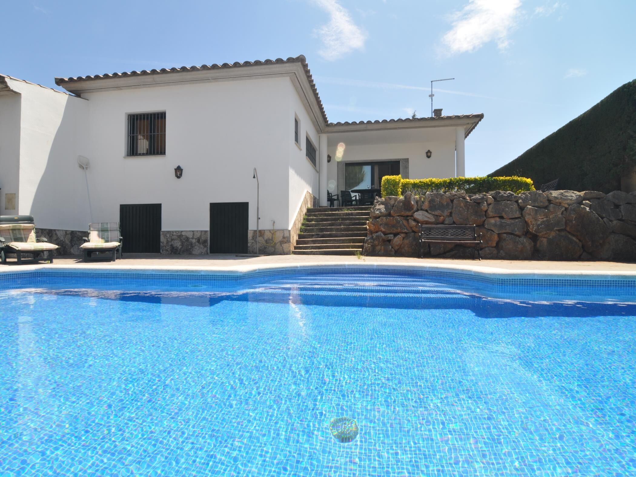 A swimming pool with a white building in the background, a stone wall, and a staircase leading to the building.