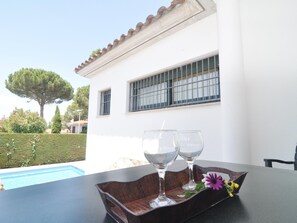 Uma mesa com duas taças de vinho e um tabuleiro decorativo, um edifício branco com janelas protegidas por grades, e uma piscina ao fundo.