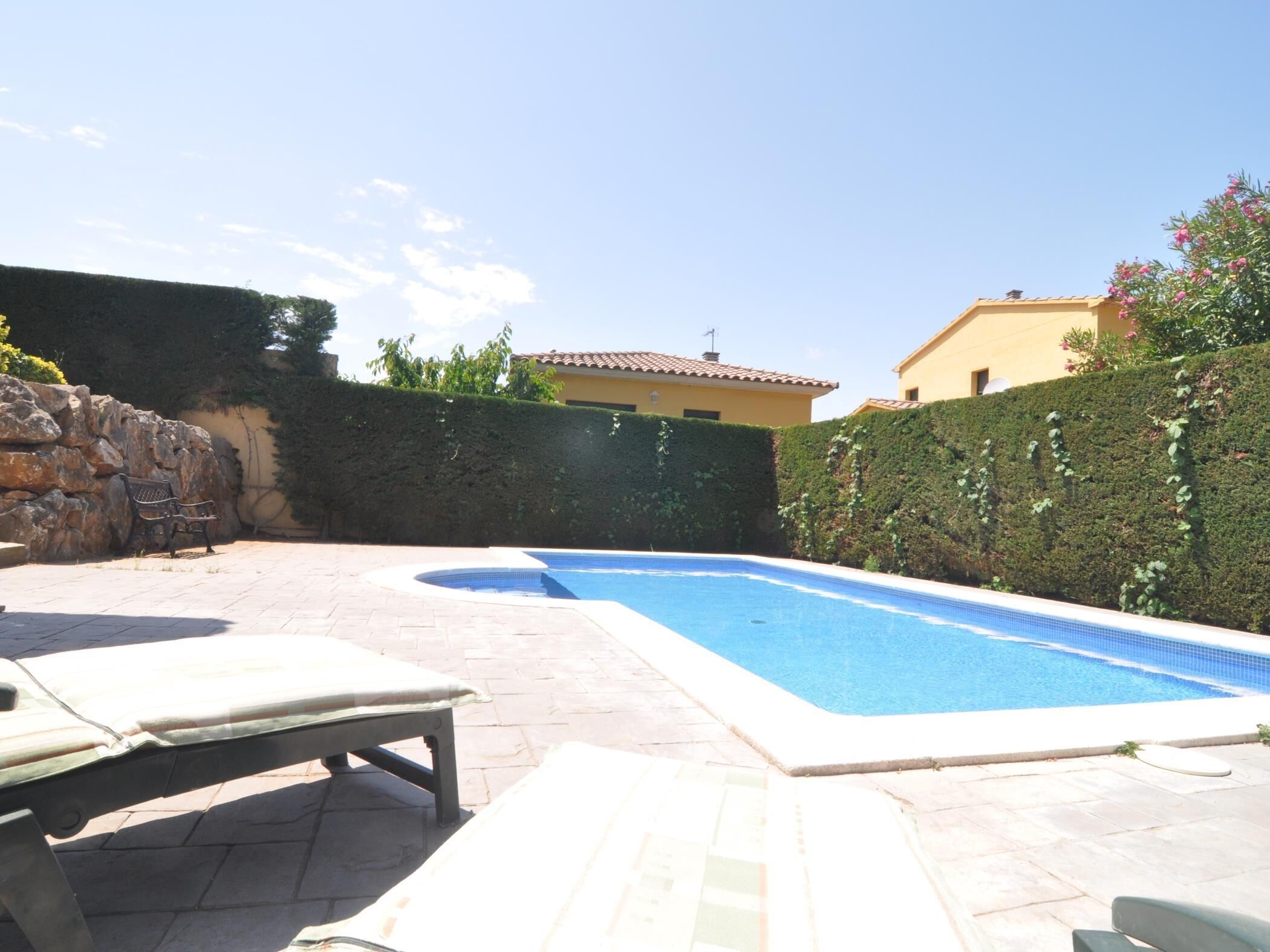 A swimming pool with a tiled edge, surrounded by a stone wall and a hedge. There are lounge chairs and a bench near the pool.