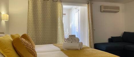 A hotel room with a bed, yellow pillows, a dark sofa, and a window with curtains.