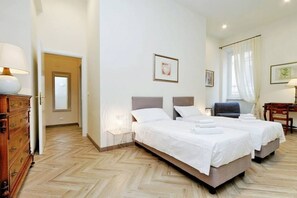 Uma cama de casal com roupa de cama branca, uma cômoda de madeira e uma cadeira em quarto de hotel com piso em padrão herringbone.