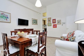 Un comedor con una mesa de madera dispuesta para cuatro, un televisor montado en la pared y un sofá blanco con cojines de colores.