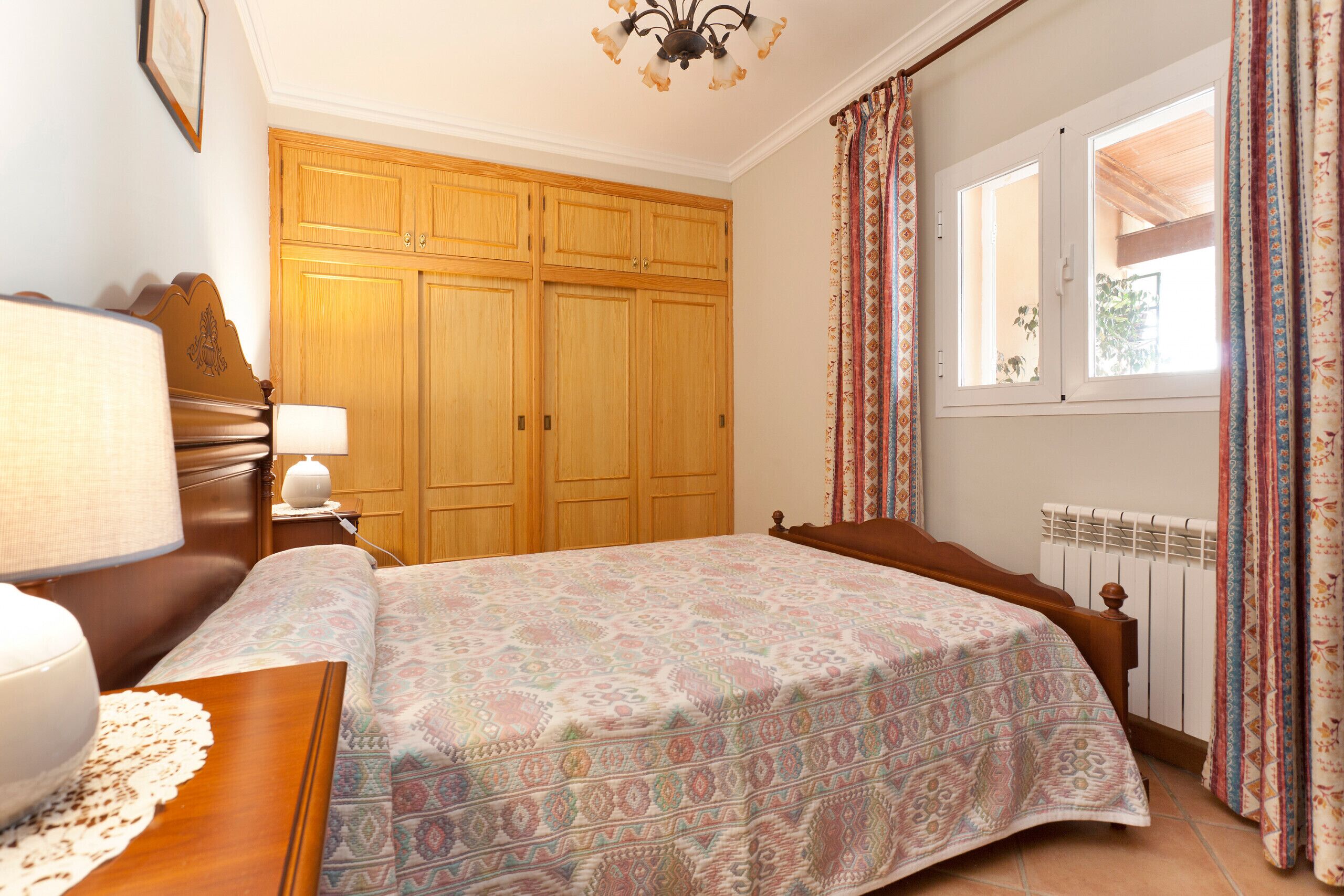 A bedroom with a bed, wooden wardrobe, bedside table with lamp, and a window with curtains.