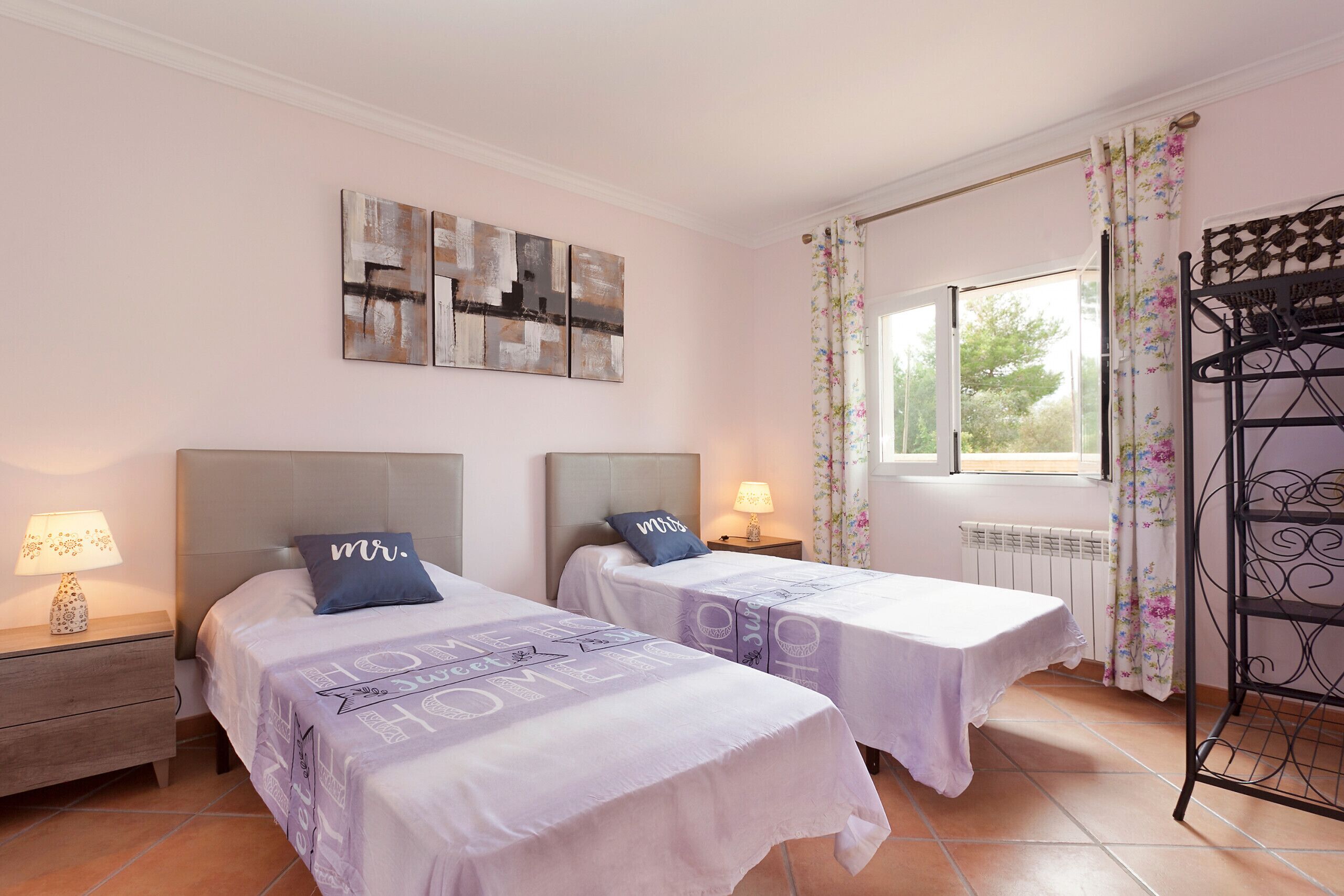A bedroom with two beds, each with a headboard, a nightstand with a lamp, and a window with curtains.