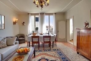 Una sala de estar con sofá, mesa de comedor, sillas, candelabro y vistas amplias por grandes ventanas.