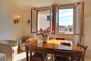 A dining area with a wooden table, chairs, a vase of flowers, and a view of buildings through open windows. - Cannaregio grande - Apartment for 7 people in Venecia (Venice)