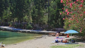 A beach with a stone wall, clear water, and a person sunbathing under a blue umbrella.